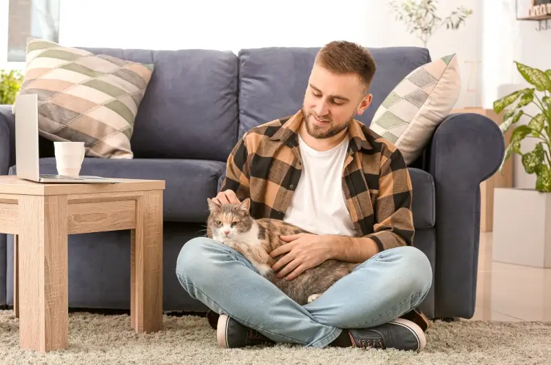 Man sitting on the floor, leaning against a couch with a tabby cat on his lap